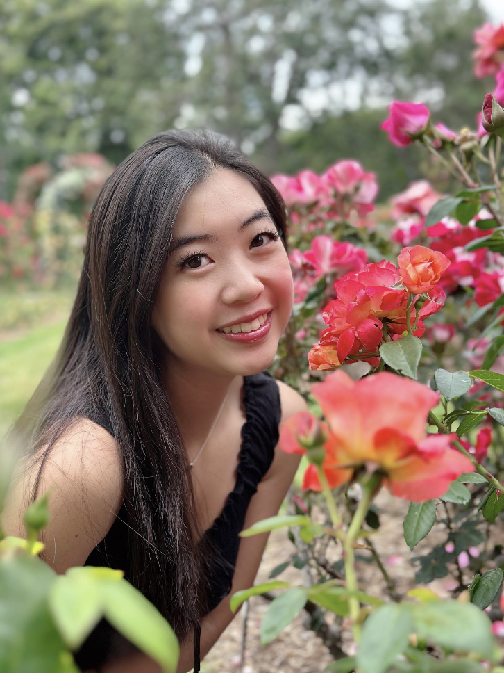 A close-up of Meileen Taw, an Asian female with black hair and brown eyes, wearing a black sleeveless top, smiling next to pink and red flowers in an outdoor garden at the Huntington Library.