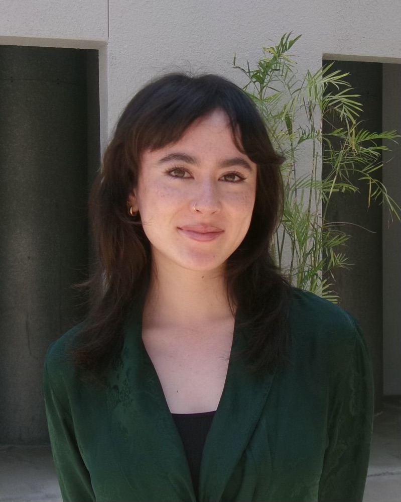 Photo of Riley Masterson from the chest up smiling at the camera. She is a light-skinned person with medium-length brown hair wearing a dark green top.