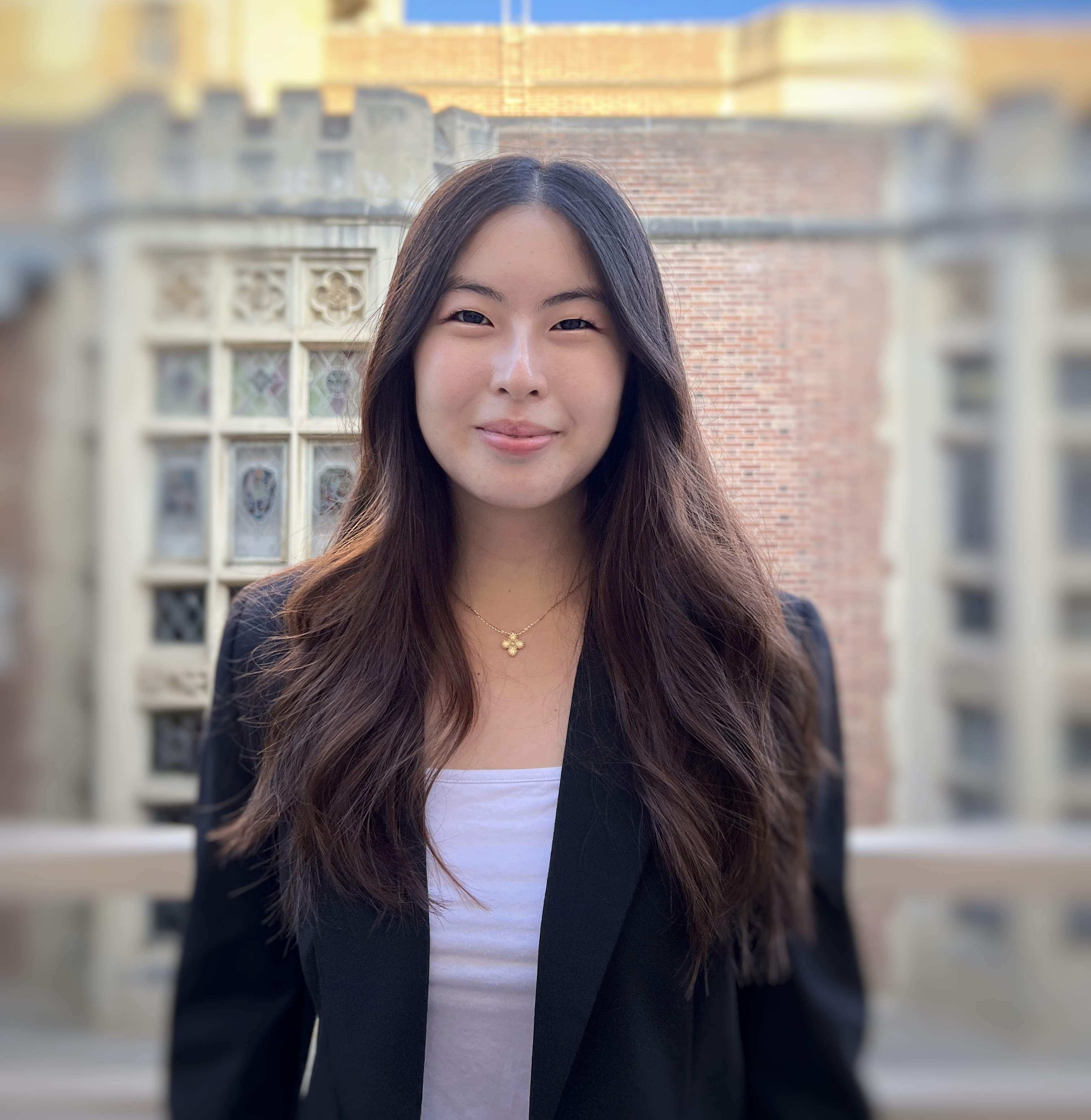 Tammy, an Asian woman with brown hair, dons a white top and black blazer as she smiles at the camera, in front of a red brick building.