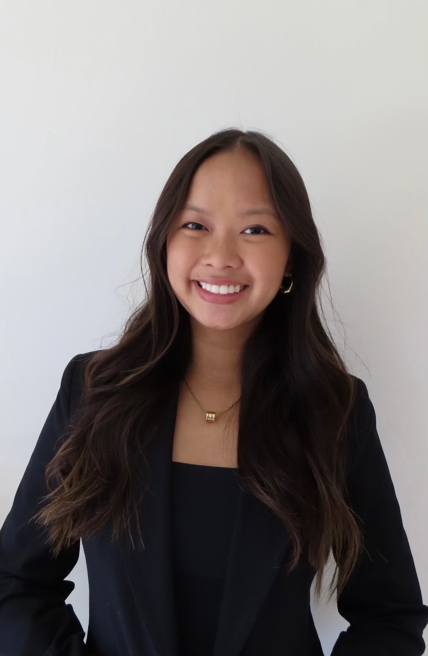 A medium shot of Tara Lam. She is an Asian woman with long black hair. She is wearing a black blazer and shirt with gold earrings and necklace. She is smiling towards the camera and her hair is curled. The background is plain white.