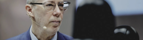 Jack Bobo wears a blue suit and glasses. He looks to the side as he listens to the speaker.