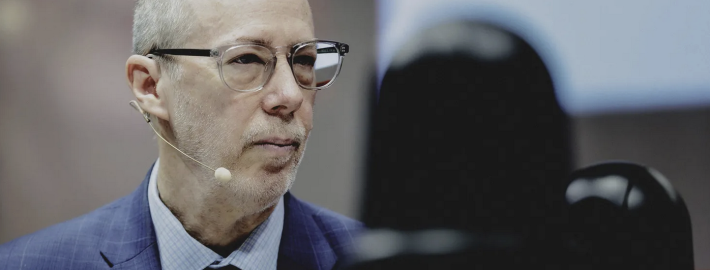 Jack Bobo wears a blue suit and glasses. He looks to the side as he listens to the speaker.