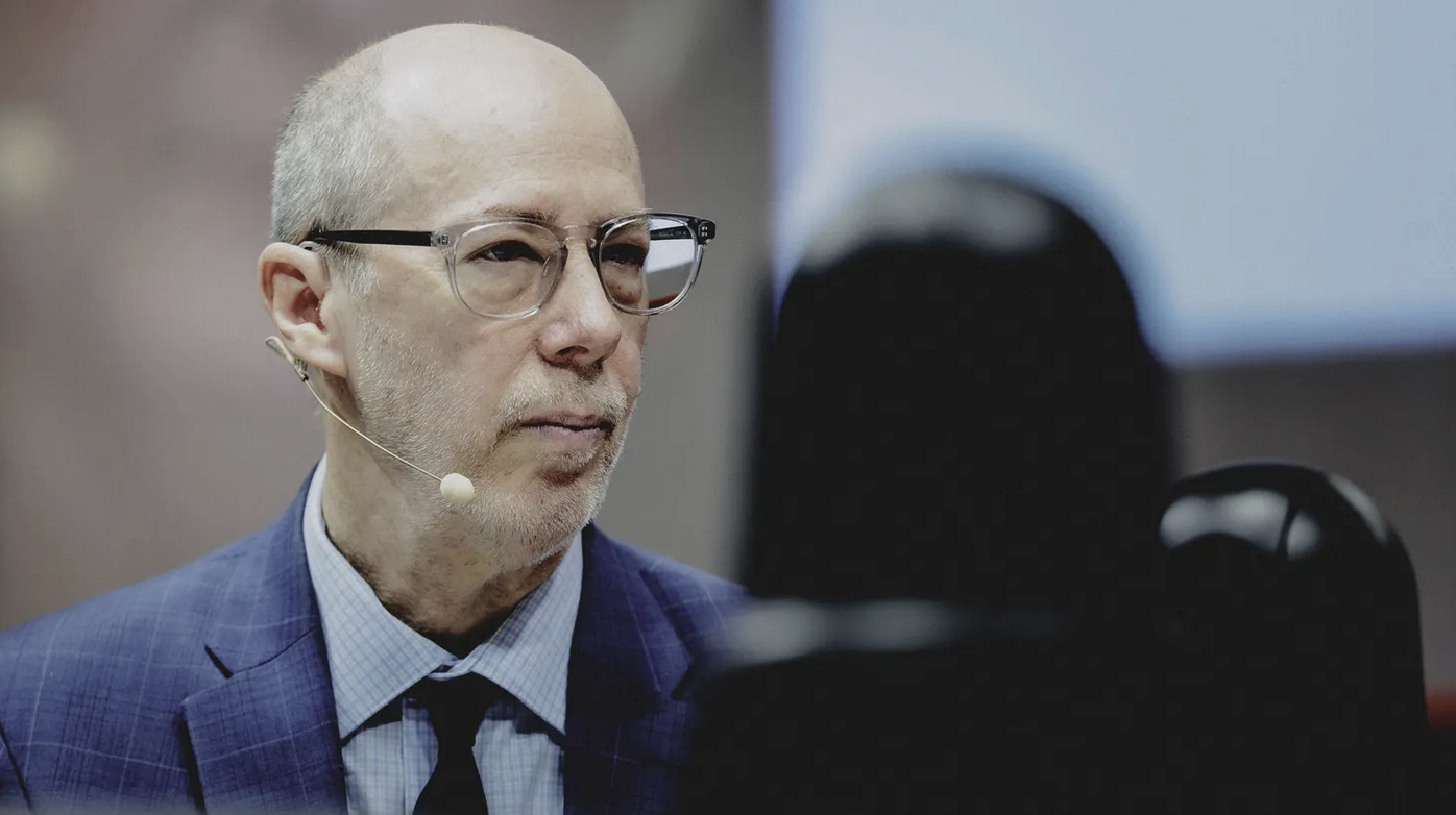 Jack Bobo wears a blue suit and glasses. He looks to the side as he listens to the speaker.