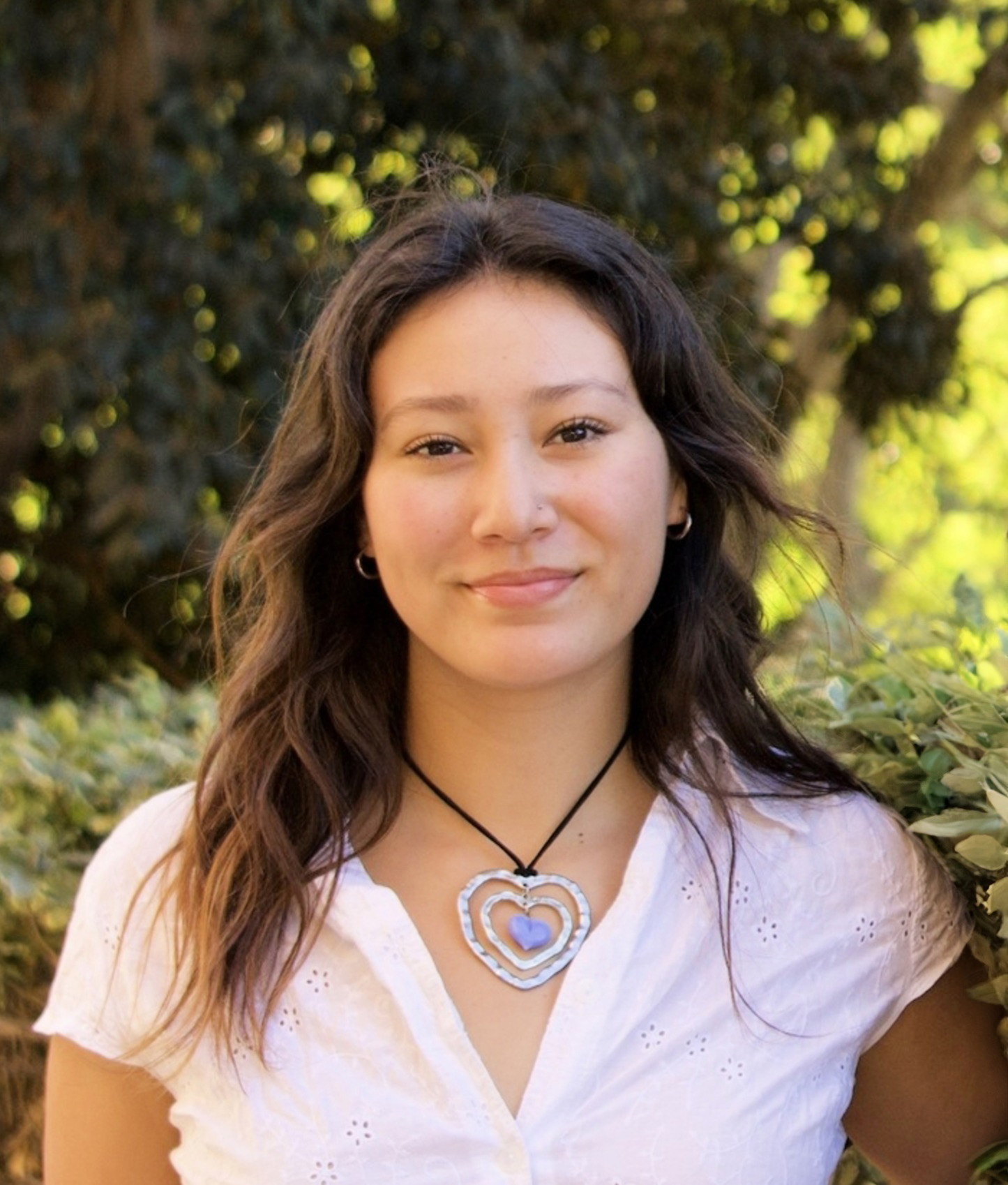 Photo of Isabel Rubin-Saika. Isabel is wearing a white V-neck button-up top and has a large heart-shaped necklace. She has medium-length brown hair. She is smiling at the camera. She is standing in front of green foliage, and there are trees in the background.