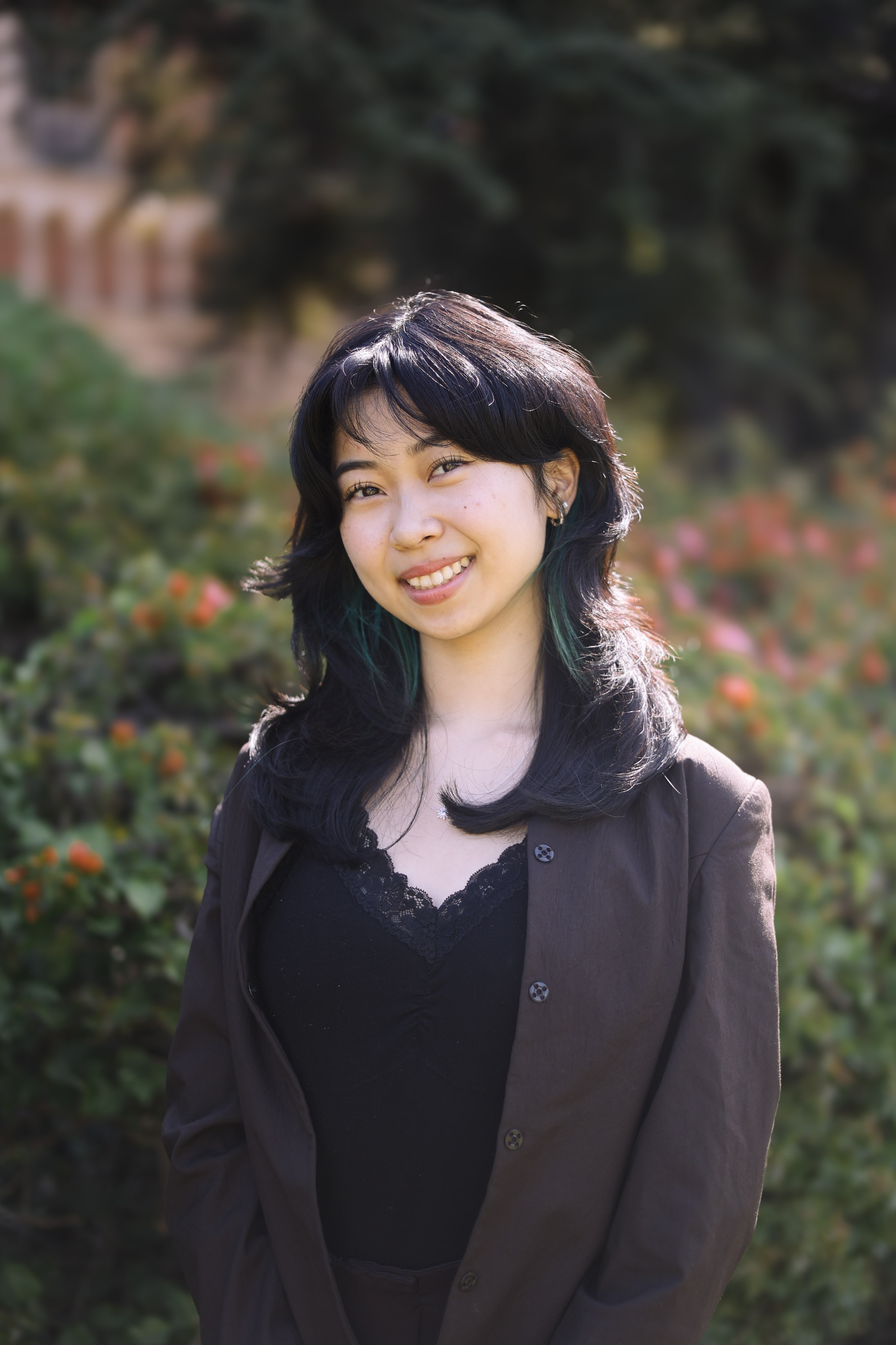Photo of Ingrid Leng. She has medium length black hair and wears a brown blazer over a black dress top. Ingrid is smiling and standing in front of a bush of flowers.